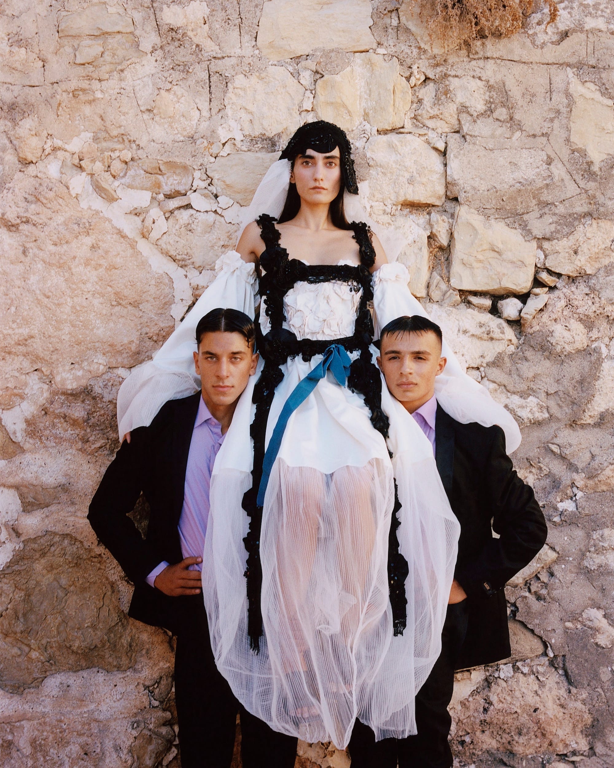 Photograph of three people (two male, one female) in front of a stone wall. all figures are dressed up festively.