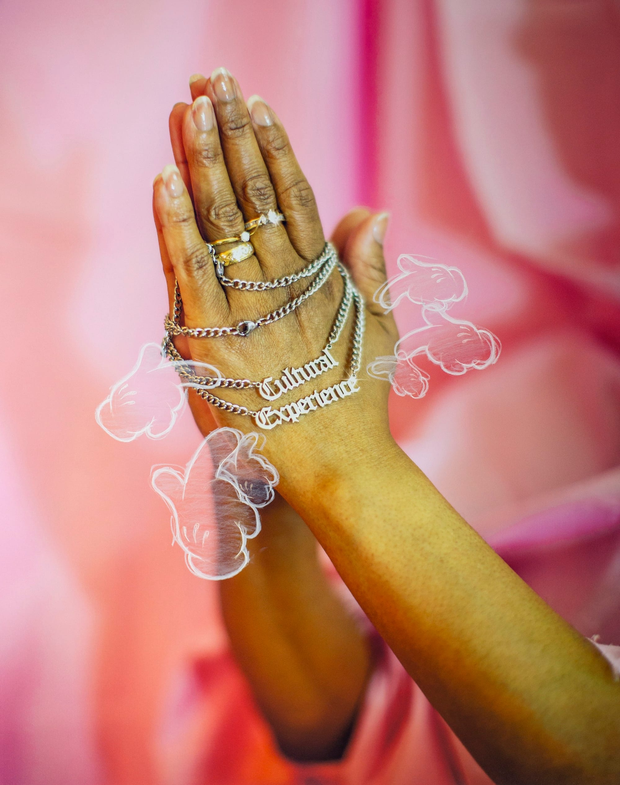 Photo of a pair of hands with rings in prayer gesture, holding a chain that spells 'Cultural Experience', in front of a pink silky background. © André Ramos-Woodard