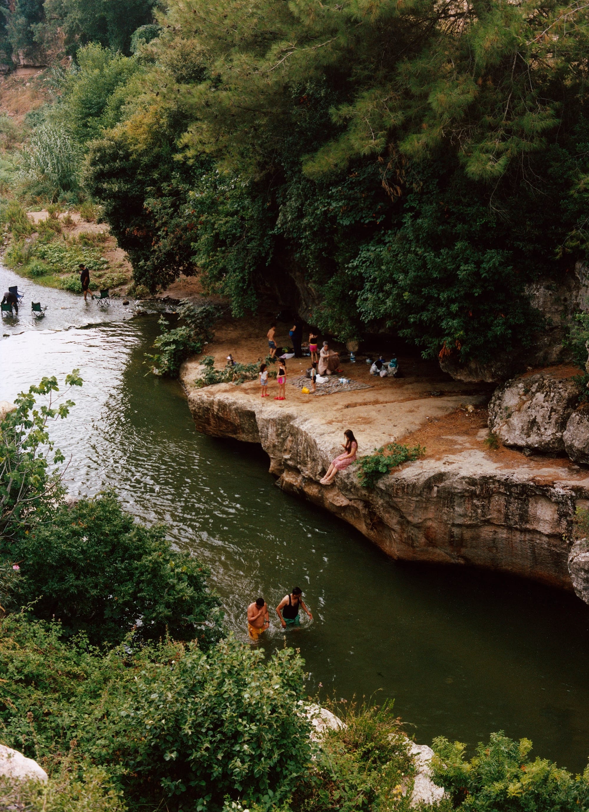 Photograph of a natural landscape from above: a river with bathing people, its shores lined with rocks and trees. © Olgaç Bozalp