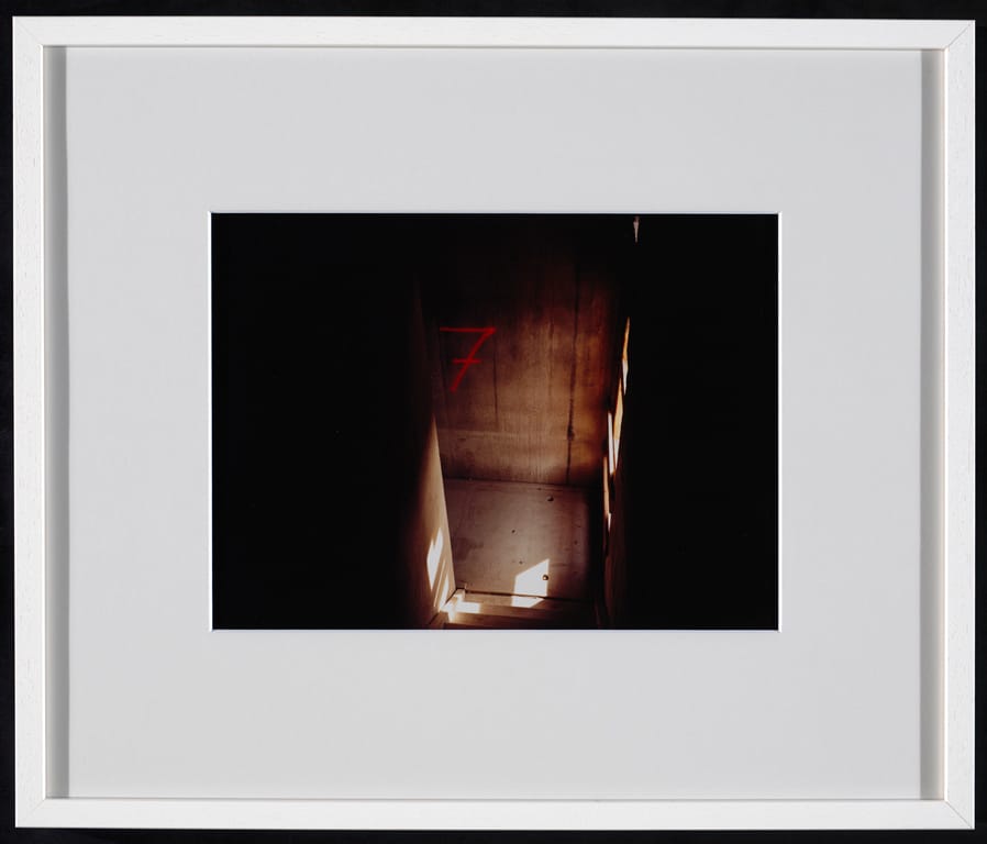 View down in a bare concrete stairwell, showing a red painted 7 on the wall. Photograph by Maarten van Schaik